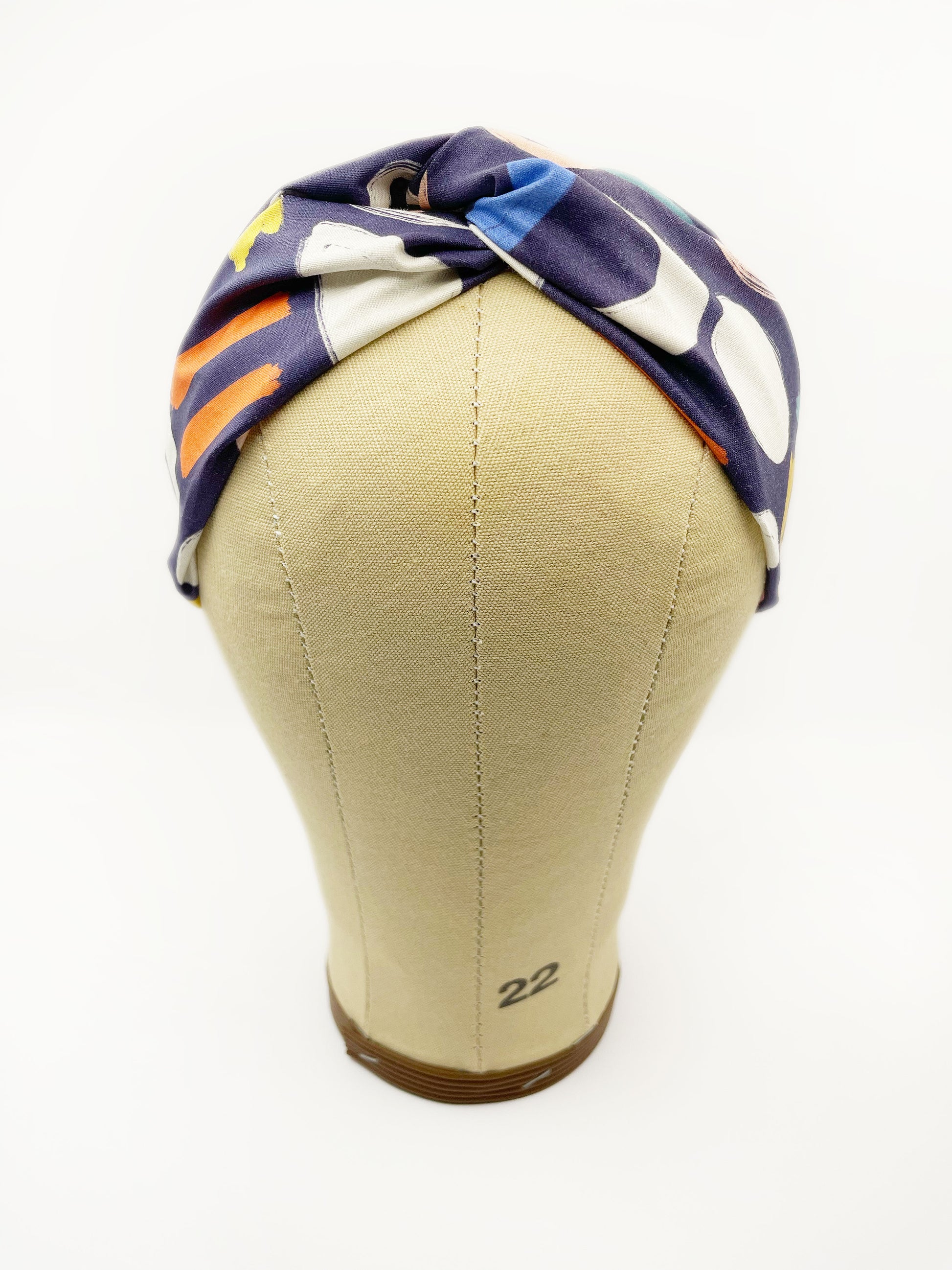 A navy blue twist headband with a colorful abstract print featuring various doodles. The headband is displayed on a mannequin head.