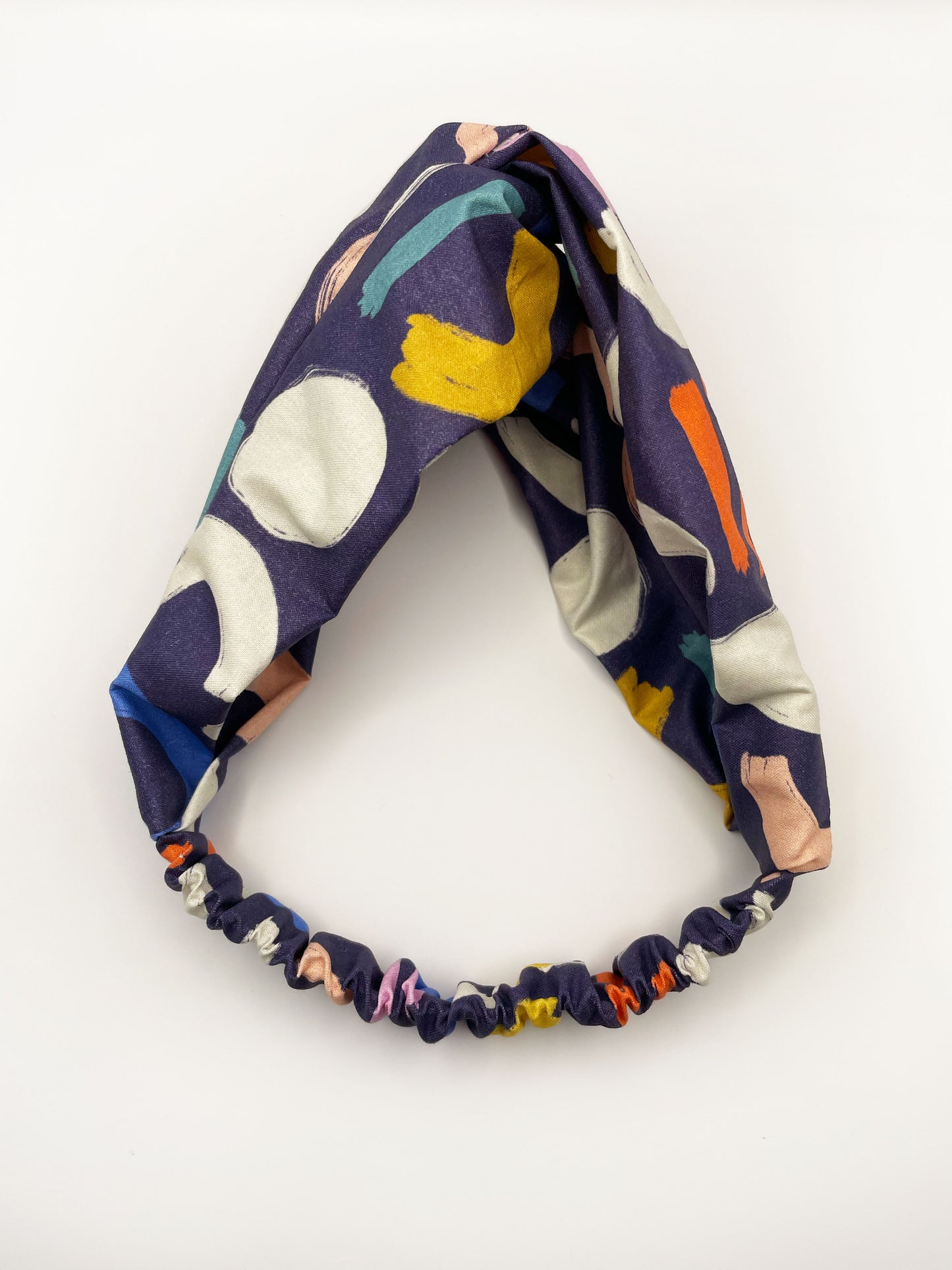 A navy blue twist headband with a colorful abstract print featuring various doodles. The elastic band in the back provides support without slipping.