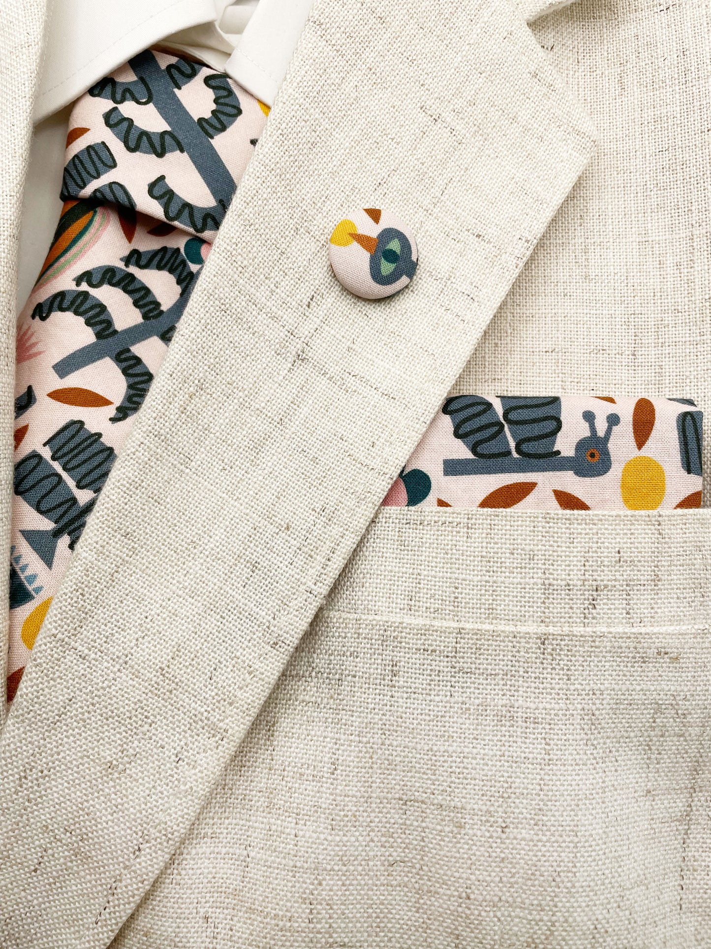 A necktie featuring a bold nature-inspired print of bugs, plants, suns, and rainbows. The color palette includes muted mustard, gentle gray, and soft pastels. The necktie is with a matching pocket square and lapel pin.