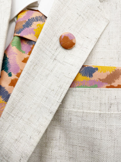 A necktie crafted from high-quality cotton with an abstract design inspired by the rolling hills and earthy tones of the desert.  The necktie is shown with a matching pocket square and lapel pin.