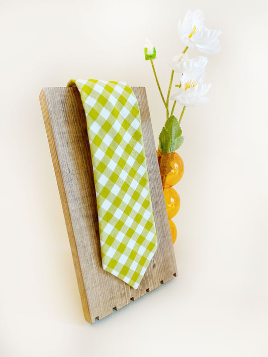 A cotton necktie with a green and white gingham plaid print. The necktie is on a wooden display next to an orange bubble vase containing white artificial flowers.