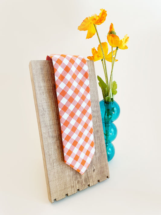 A cotton necktie with an orange and white gingham plaid print. The necktie is displayed on a wooden stand next to a blue bubble vase containing orange artificial flowers.