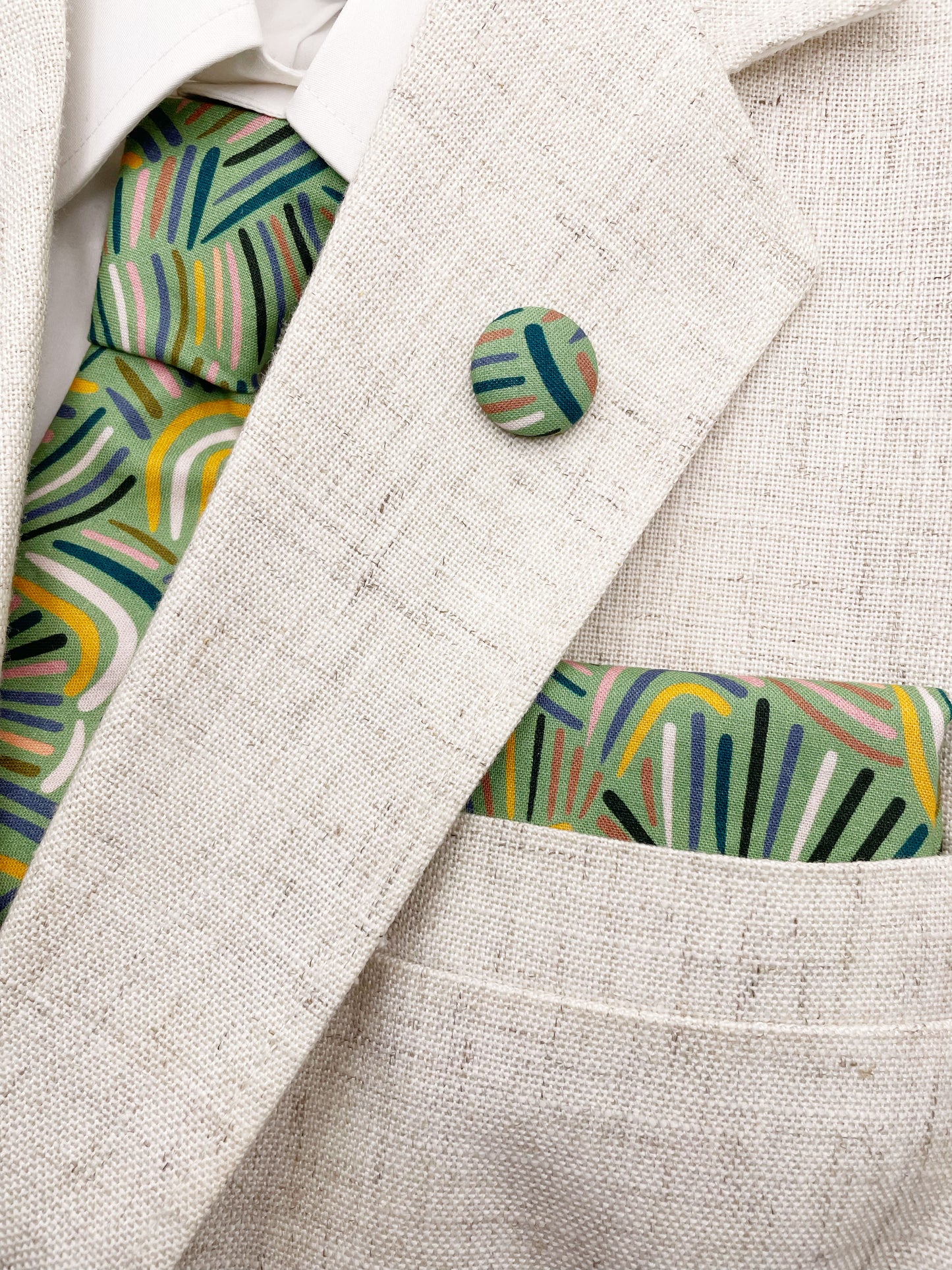 A necktie made from high-quality cotton with an abstract design inspired by desert rain patterns. The necktie is shown with a matching pocket square and lapel pin.