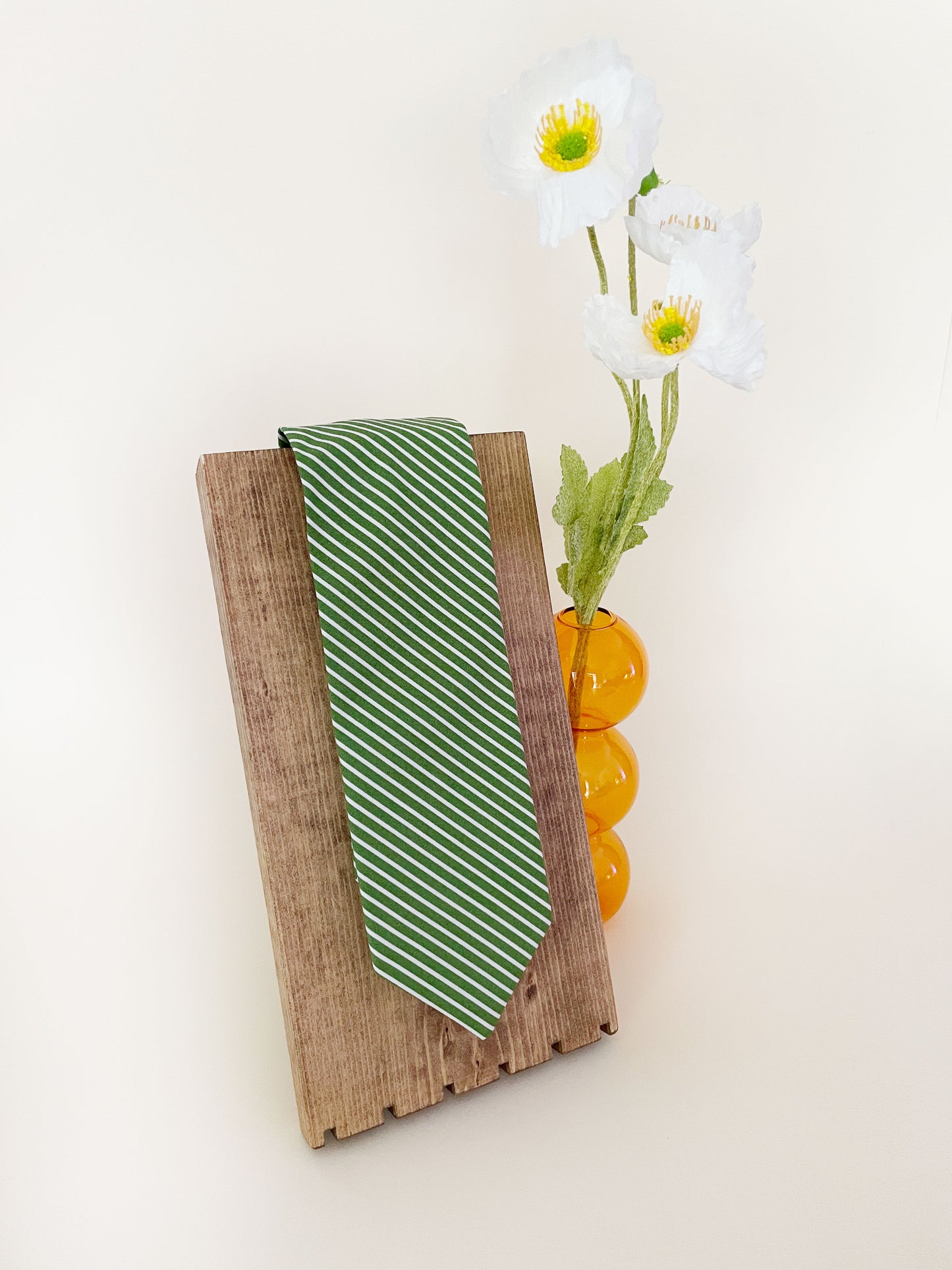 A handmade green necktie with white stripes on display next to an orange vase containing white flowers.