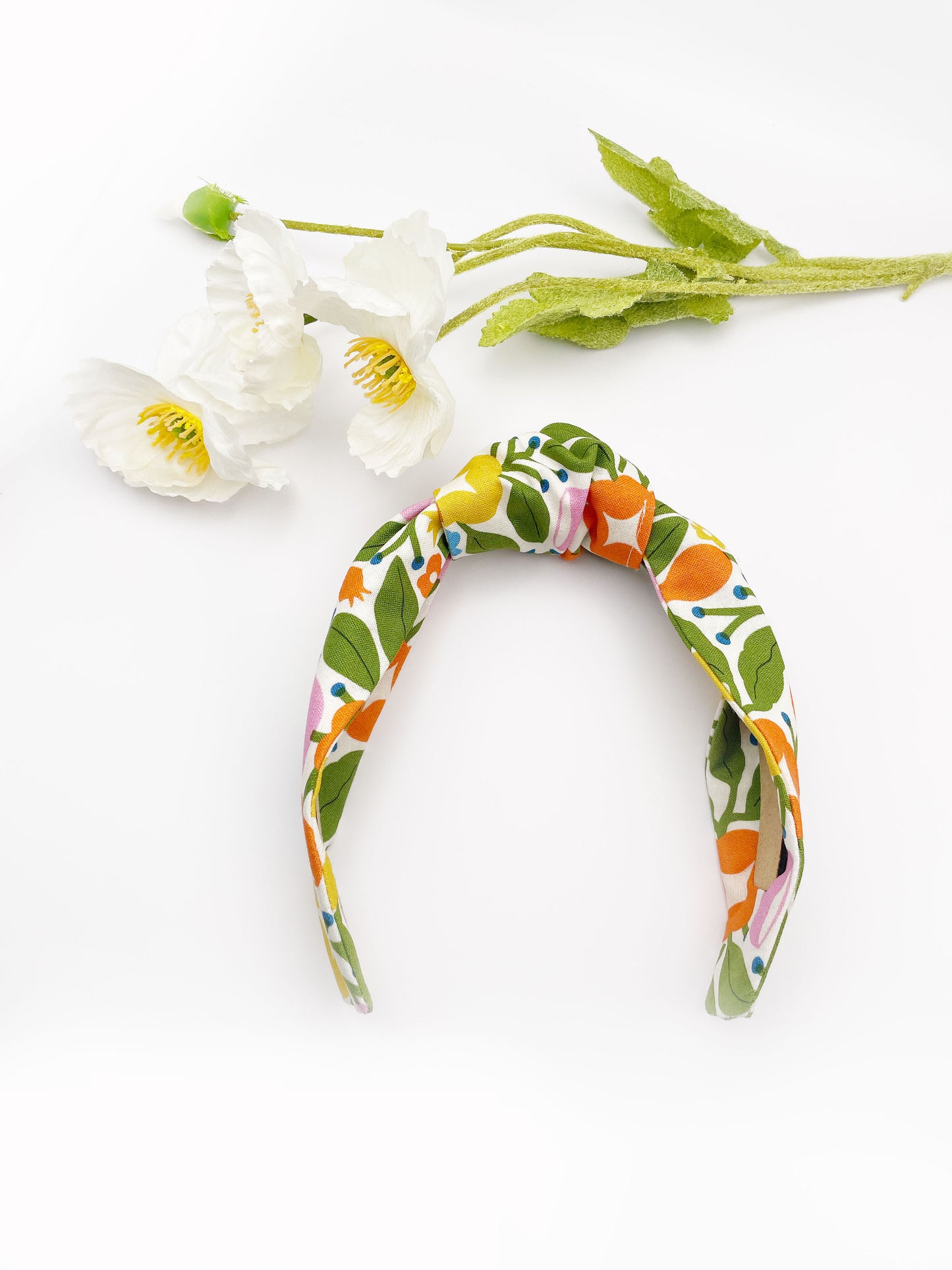 A vibrant knotted headband with bold yellow, orange, and pink flowers. The headband is next to some white artificial flowers.