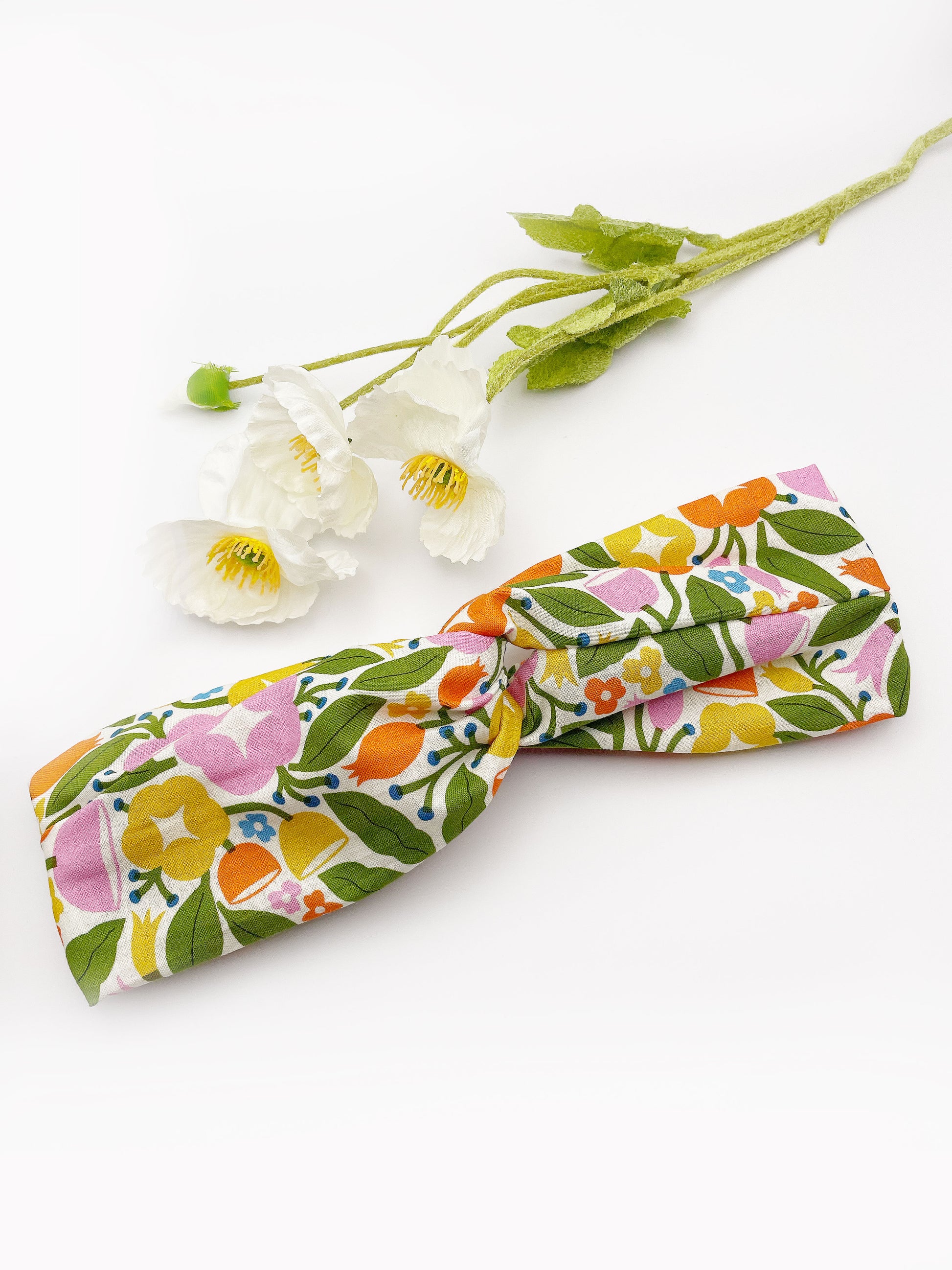 A vibrant twist headband with bold yellow, orange, and pink flowers. The headband is next to some white artificial flowers.