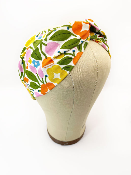 A vibrant twist headband with bold yellow, orange, and pink flowers. The headband is modeled on a mannequin head.