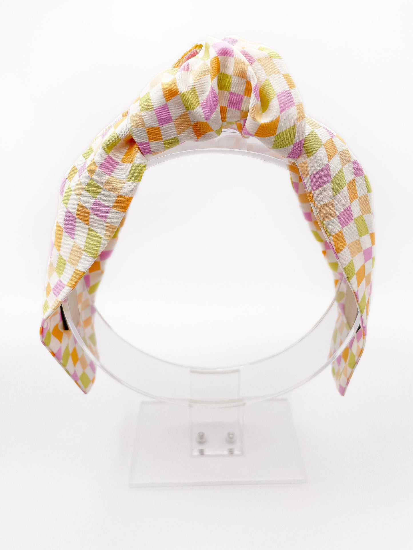 A handmade knotted headband with a pastel checkered print. The headband is displayed on an acrylic stand.