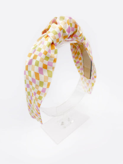 A handmade knotted headband with a pastel checkered print. The headband is displayed on an acrylic stand.