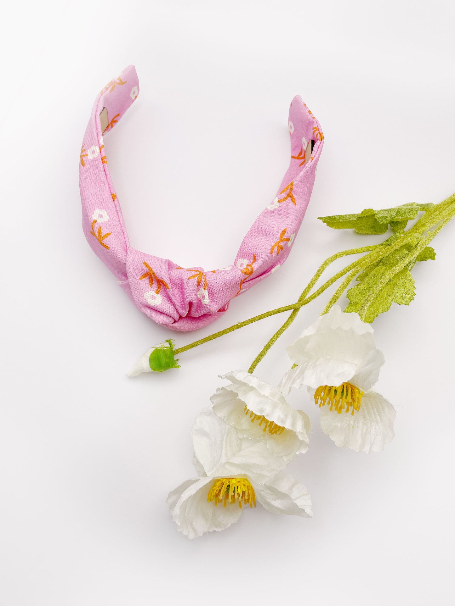 A cheerful pink knotted headband with orange-stemmed flowers. The headband is next to some white artificial flowers.