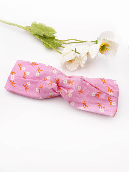 A cheerful pink twist headband with orange-stemmed flowers. The headband is next to some white artificial flowers.