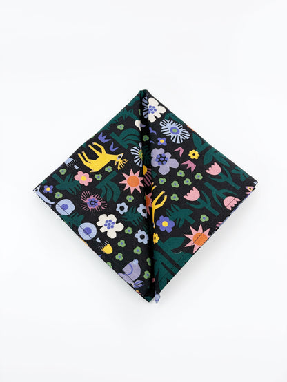 A black pocket square with a colorful abstract forest print featuring playful florals, leaves, and bright animals.