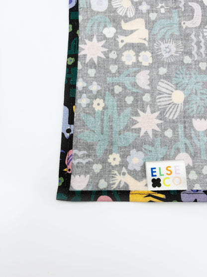 A black pocket square with a colorful abstract forest print featuring playful florals, leaves, and bright animals.