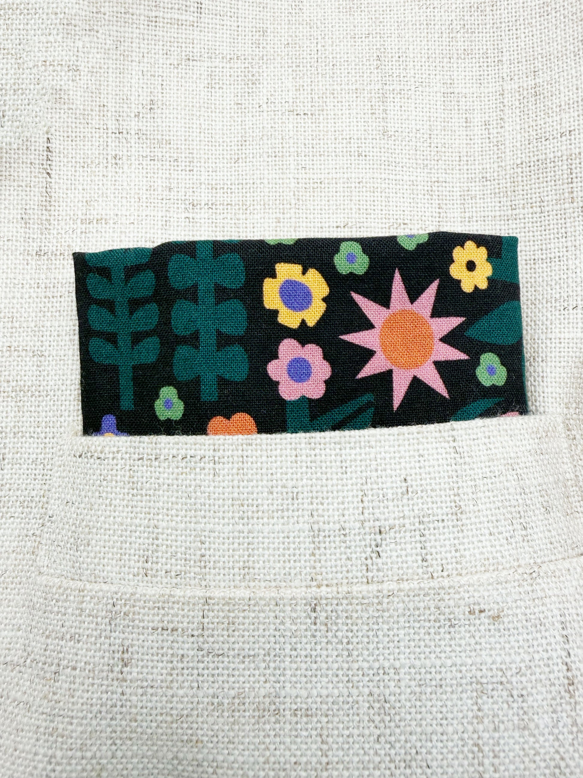 A black pocket square with a colorful abstract forest print featuring playful florals, leaves, and bright animals. The pocket square is shown in the pocket of a suit jacket.