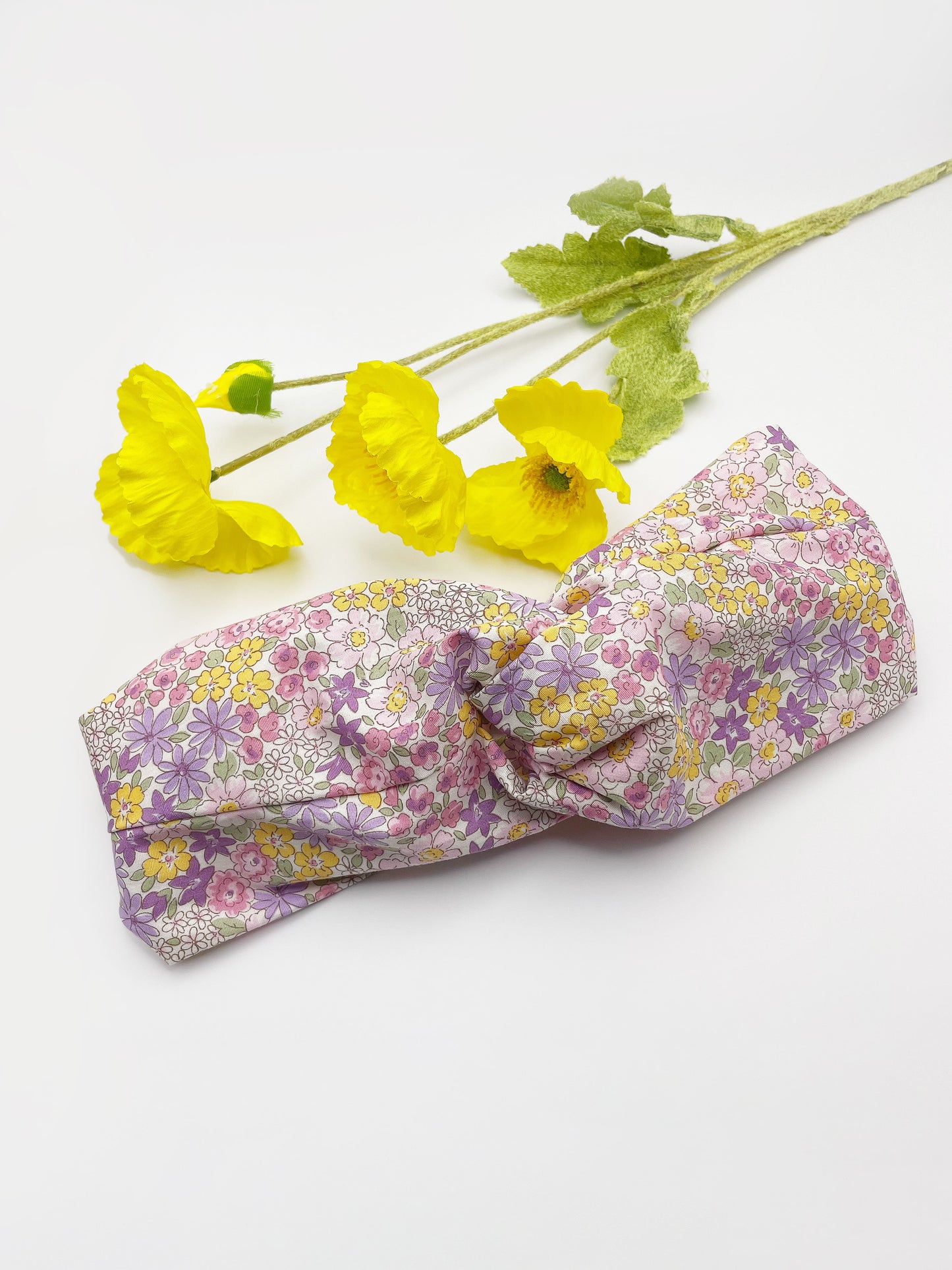 A handmade twist headband with purple flowers. The headband has an elastic band in the back. The headband is next to some yellow artificial flowers.