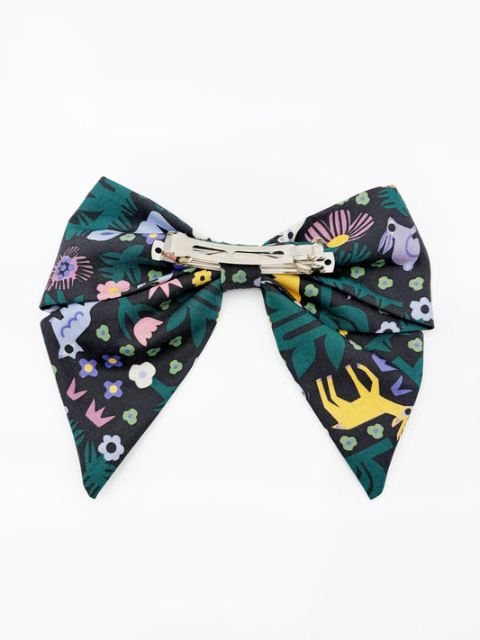 A black hair bow with a colorful abstract forest print featuring playful florals, leaves, and bright animals. The cotton fabric is attached to a sturdy barrette clip.