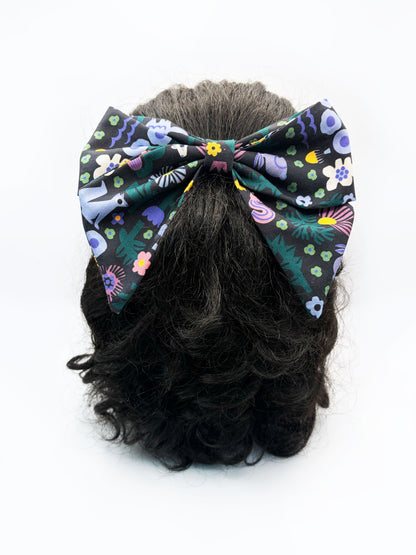A black hair bow with a colorful abstract forest print featuring playful florals, leaves, and bright animals. The bow is shown on a mannequin head.