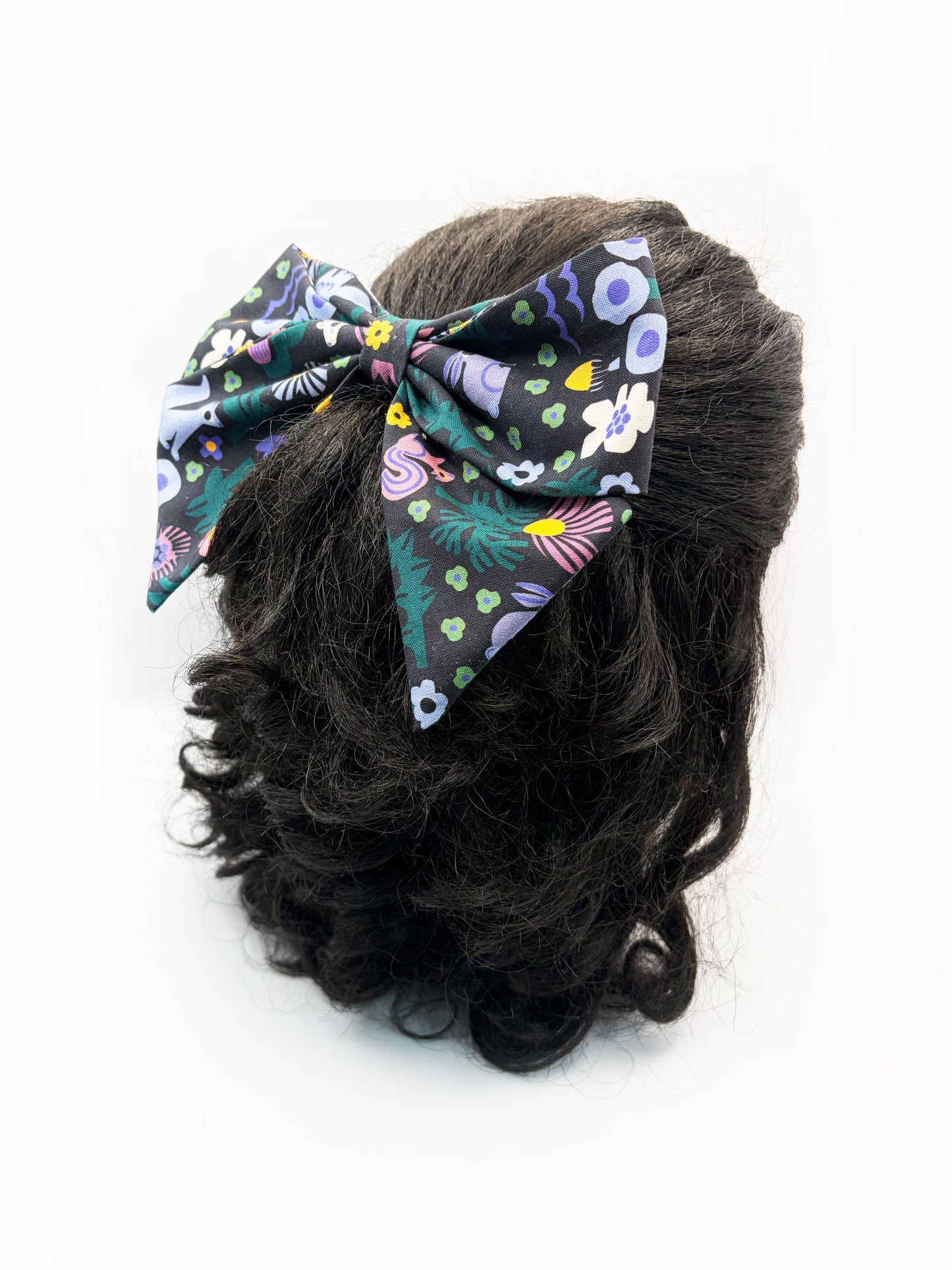 A black hair bow with a colorful abstract forest print featuring playful florals, leaves, and bright animals. The bow is shown on a mannequin head.