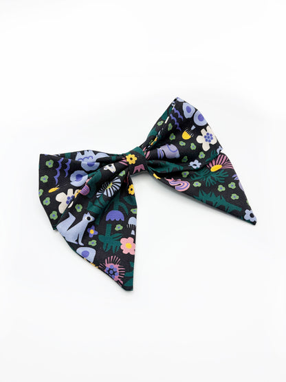 A black hair bow with a colorful abstract forest print featuring playful florals, leaves, and bright animals. 