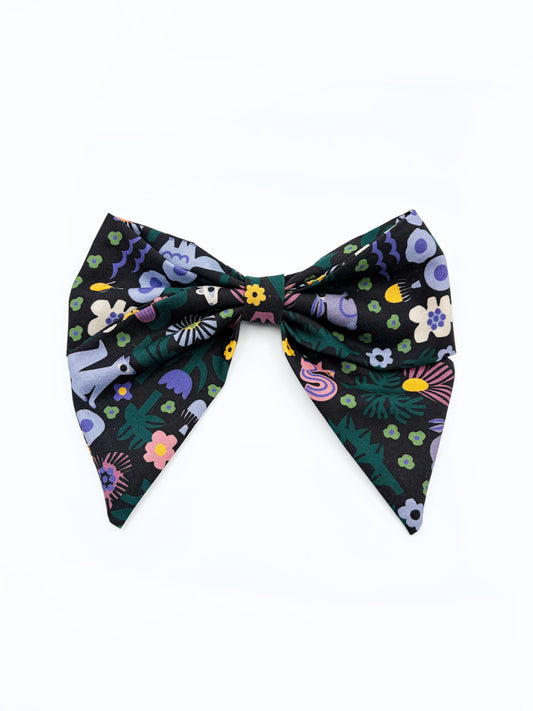 A black hair bow with a colorful abstract forest print featuring playful florals, leaves, and bright animals.