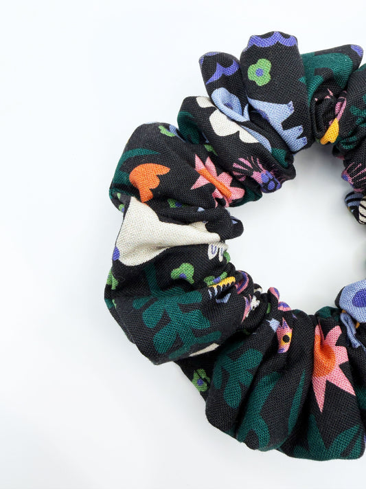 A black scrunchie with a colorful abstract forest print featuring playful florals, leaves, and bright animals.