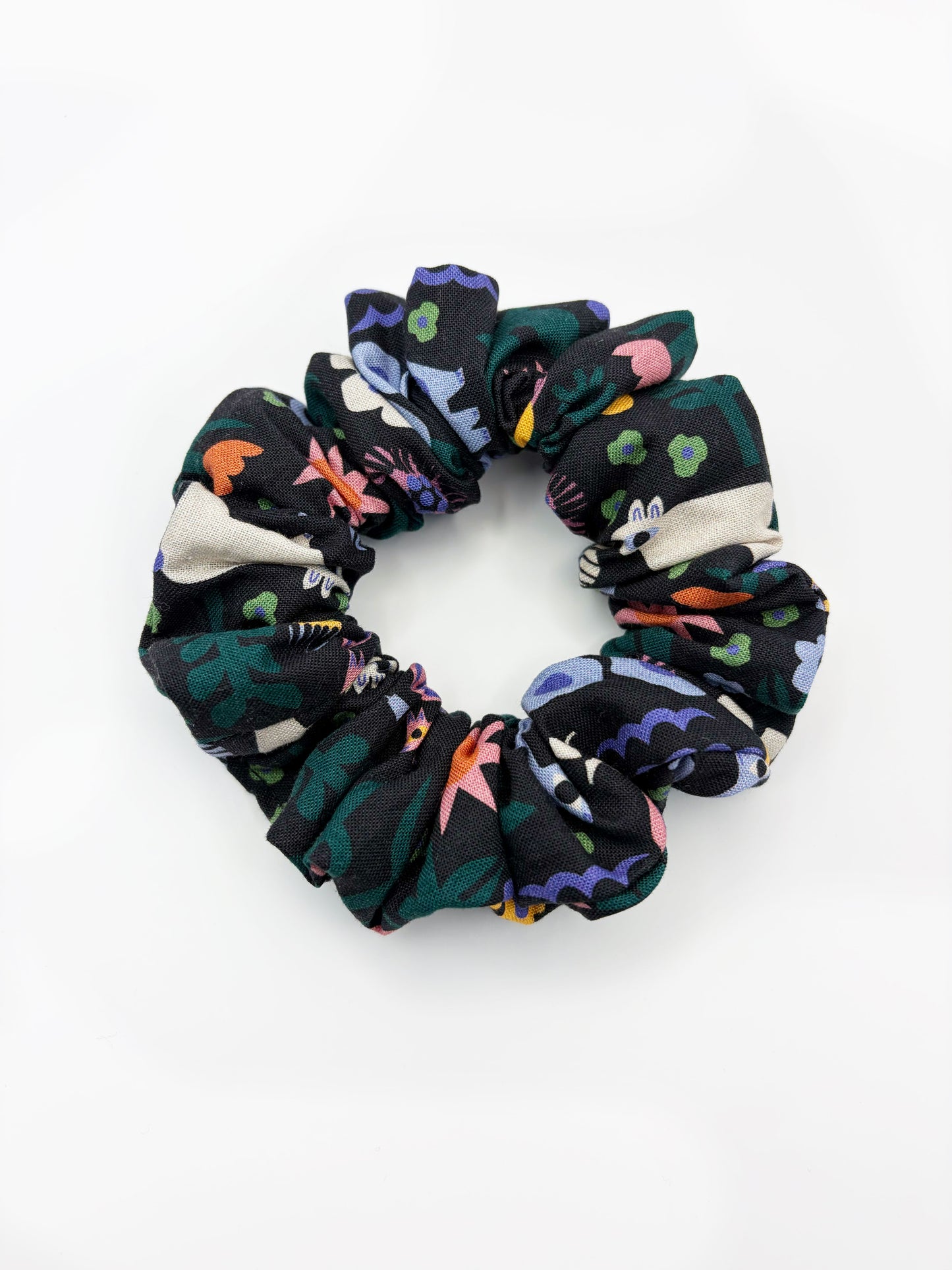 A black scrunchie with a colorful abstract forest print featuring playful florals, leaves, and bright animals.