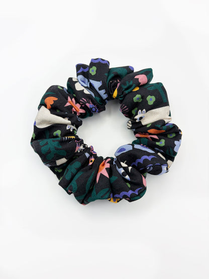 A black scrunchie with a colorful abstract forest print featuring playful florals, leaves, and bright animals.