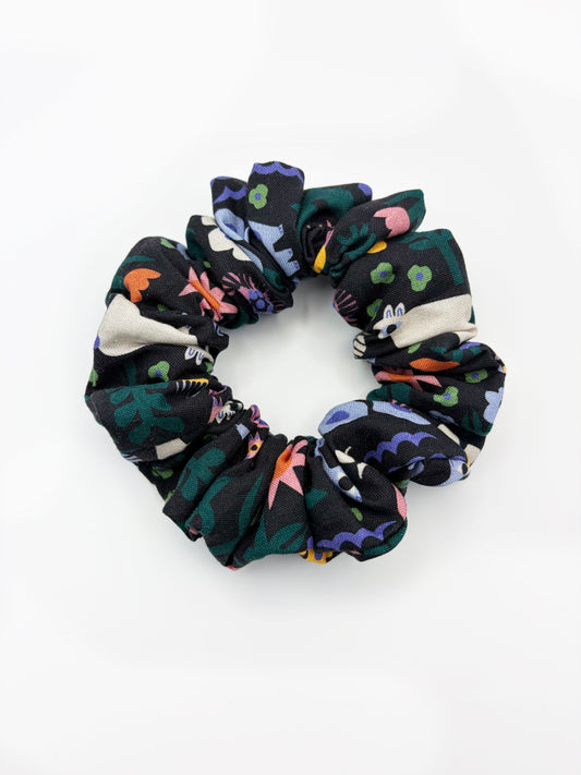 A black scrunchie with a colorful abstract forest print featuring playful florals, leaves, and bright animals.