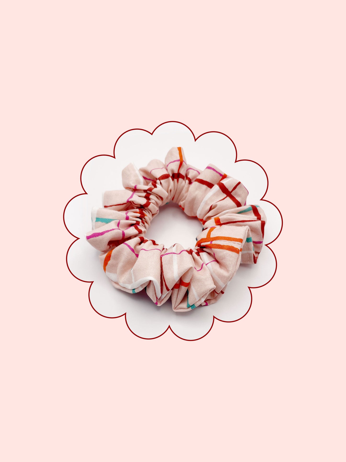 A handmade scrunchie featuring a colorful and modern plaid design with vibrant shades of pink, red, teal, and orange on a soft pastel background.