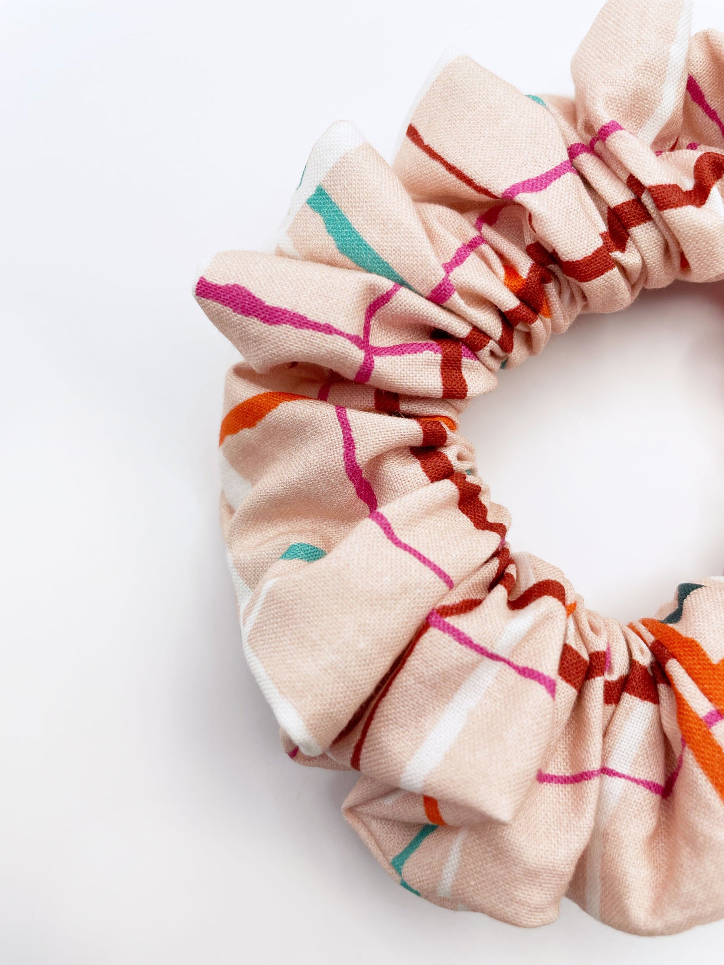 A handmade scrunchie featuring a colorful and modern plaid design with vibrant shades of pink, red, teal, and orange on a soft pastel background.