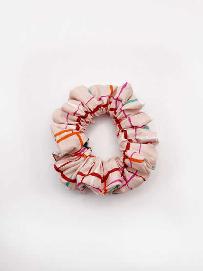 A handmade scrunchie featuring a colorful and modern plaid design with vibrant shades of pink, red, teal, and orange on a soft pastel background.