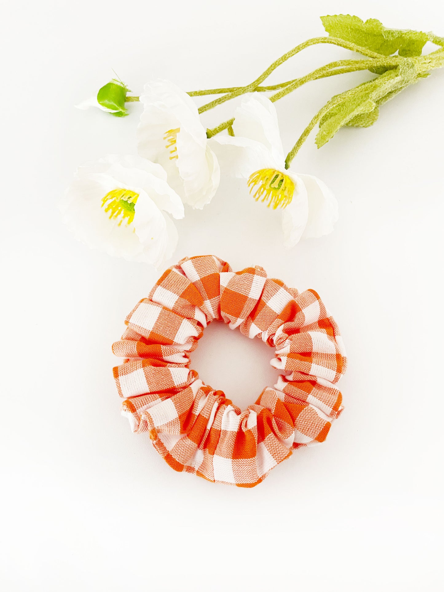 An orange and white gingham plaid scrunchie. The scrunchie is next to white artificial flowers.