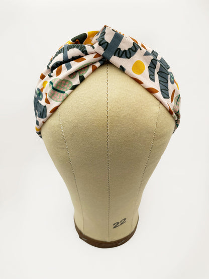 A twist headband featuring a bold nature-inspired print of bugs, plants, suns, and rainbows. The headband is shown on a mannequin head.