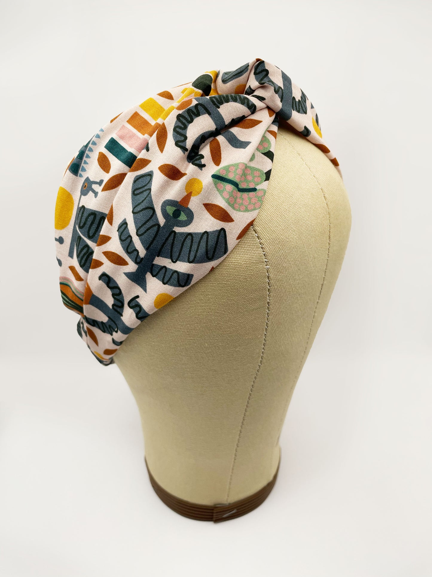 A twist headband featuring a bold nature-inspired print of bugs, plants, suns, and rainbows. The headband is shown on a mannequin head.