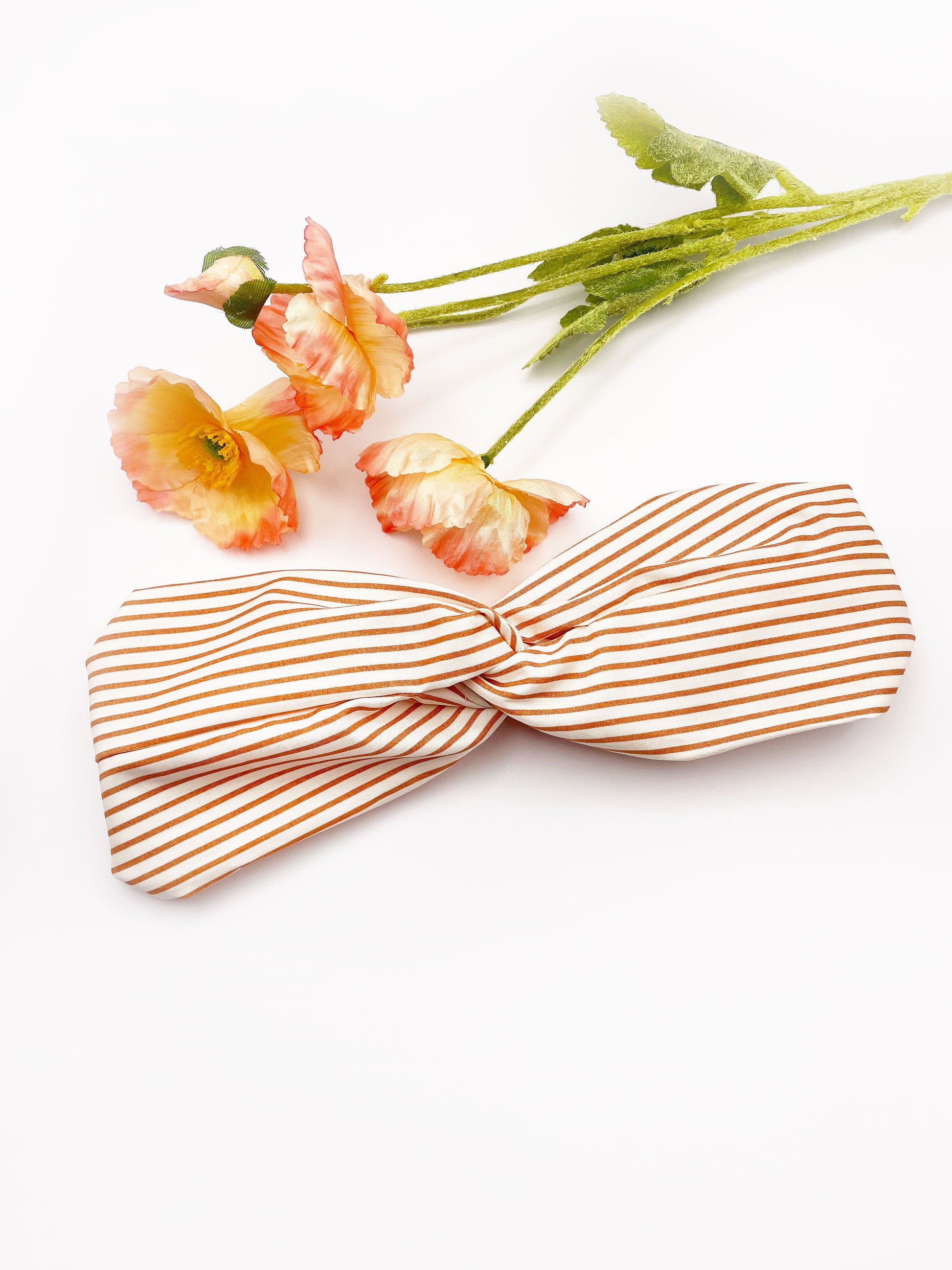 A handmade tan striped twist headband with an elastic band in the back. The headband is next to artificial flowers.