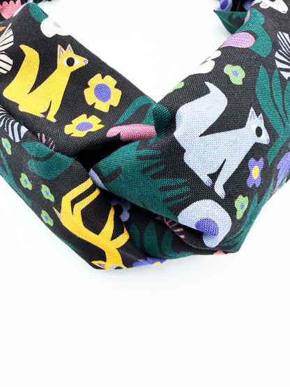 A black twist headband with a colorful abstract forest print featuring playful florals, leaves, and bright animals.