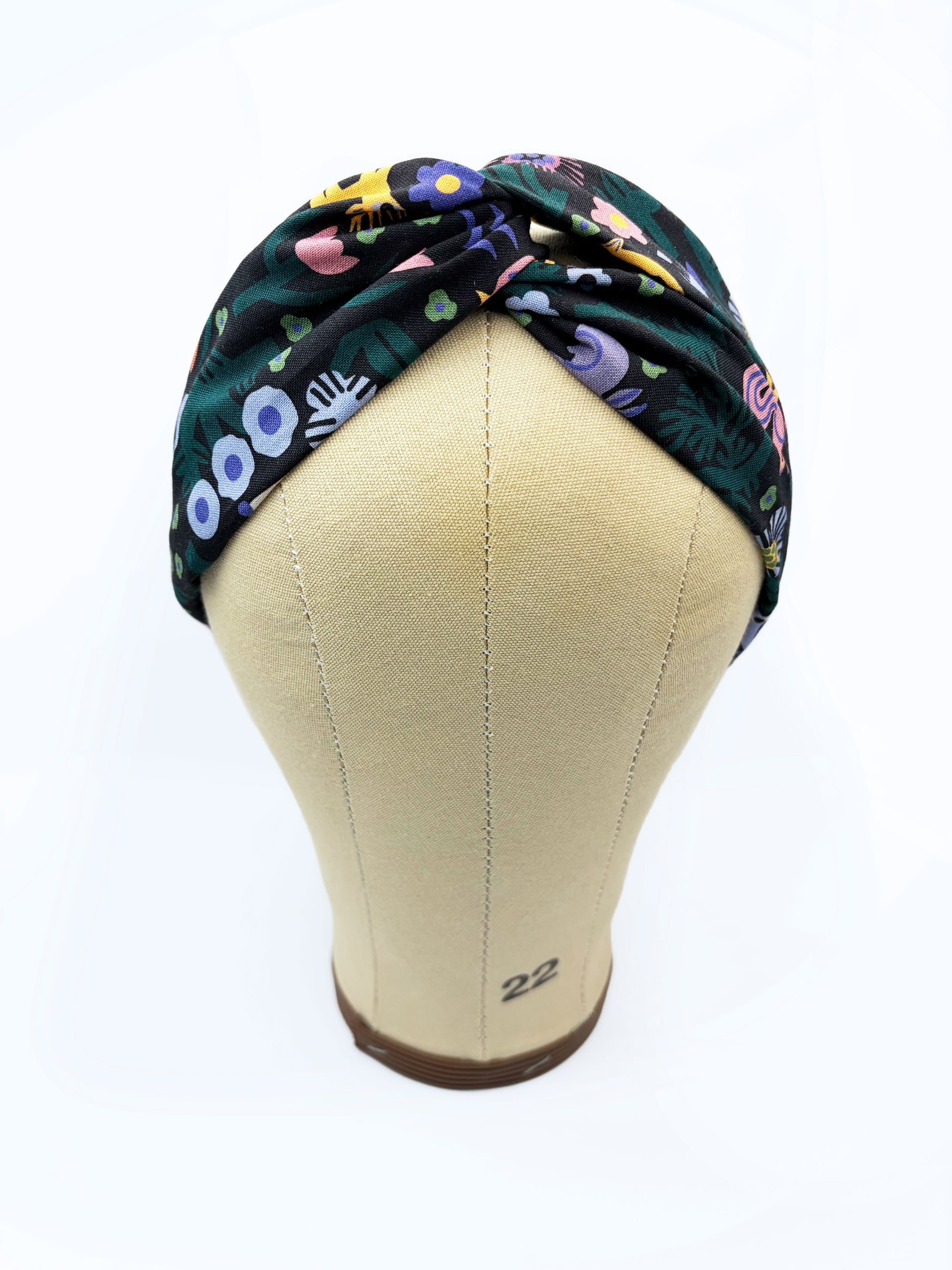 A black twist headband with a colorful abstract forest print featuring playful florals, leaves, and bright animals. The headband is shown on a mannequin head.