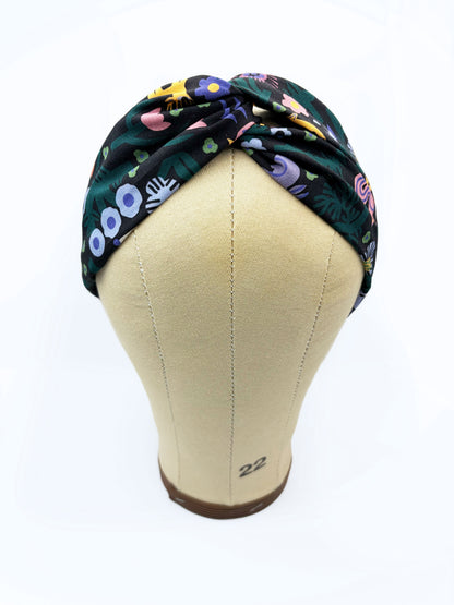 A black twist headband with a colorful abstract forest print featuring playful florals, leaves, and bright animals. The headband is shown on a mannequin head.