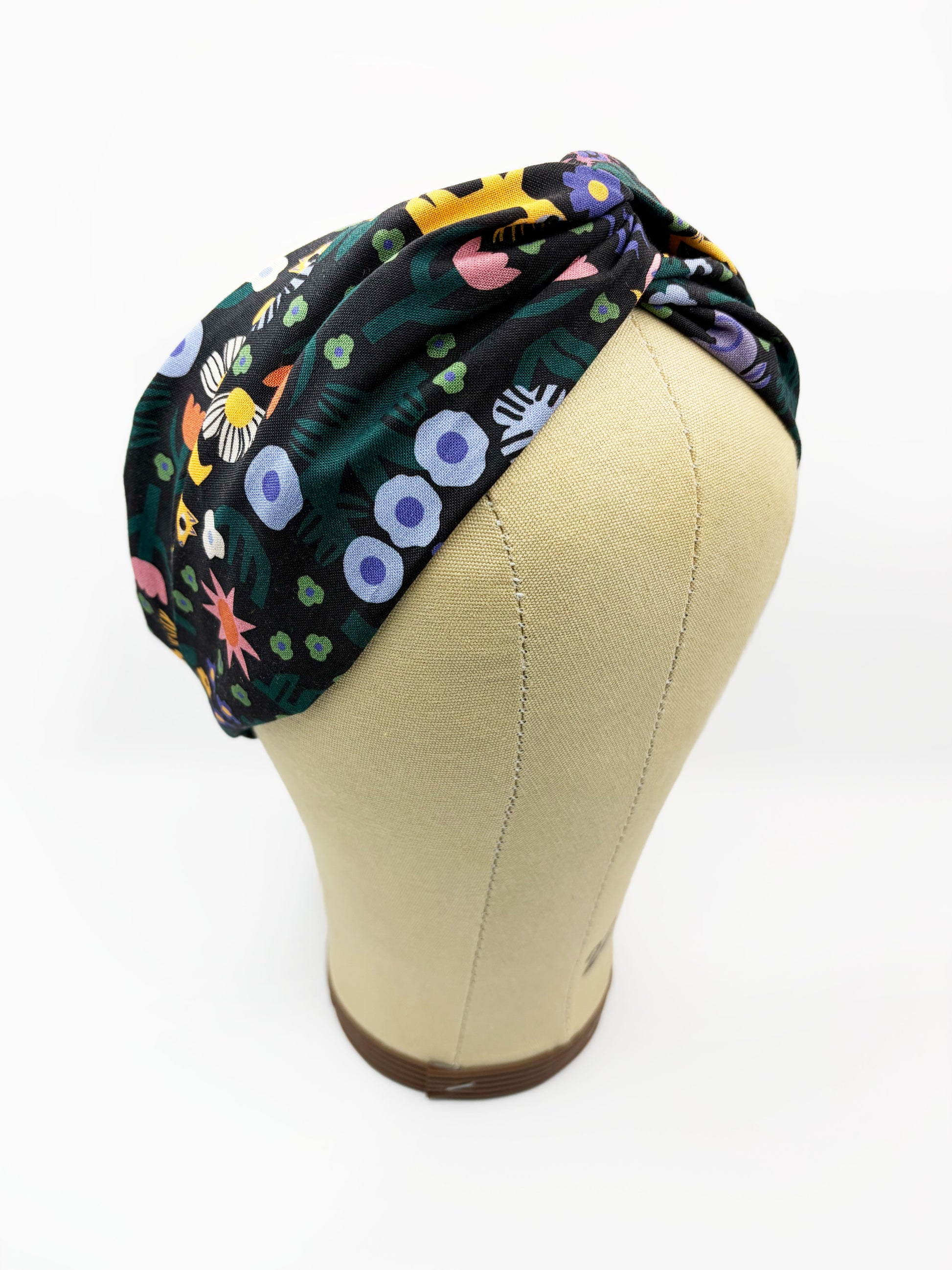 A black twist headband with a colorful abstract forest print featuring playful florals, leaves, and bright animals. The headband is shown on a mannequin head.