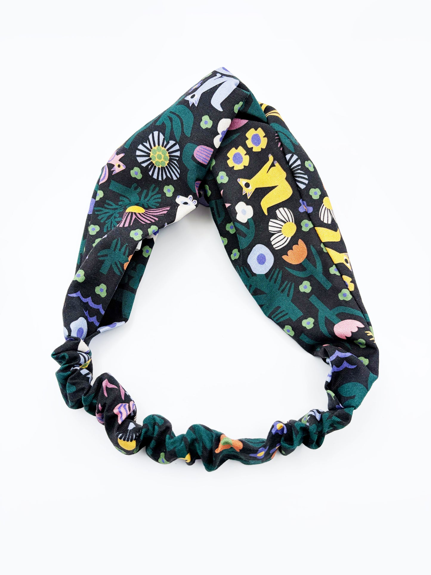 A black twist headband with a colorful abstract forest print featuring playful florals, leaves, and bright animals. The headband has an elastic band on the back.