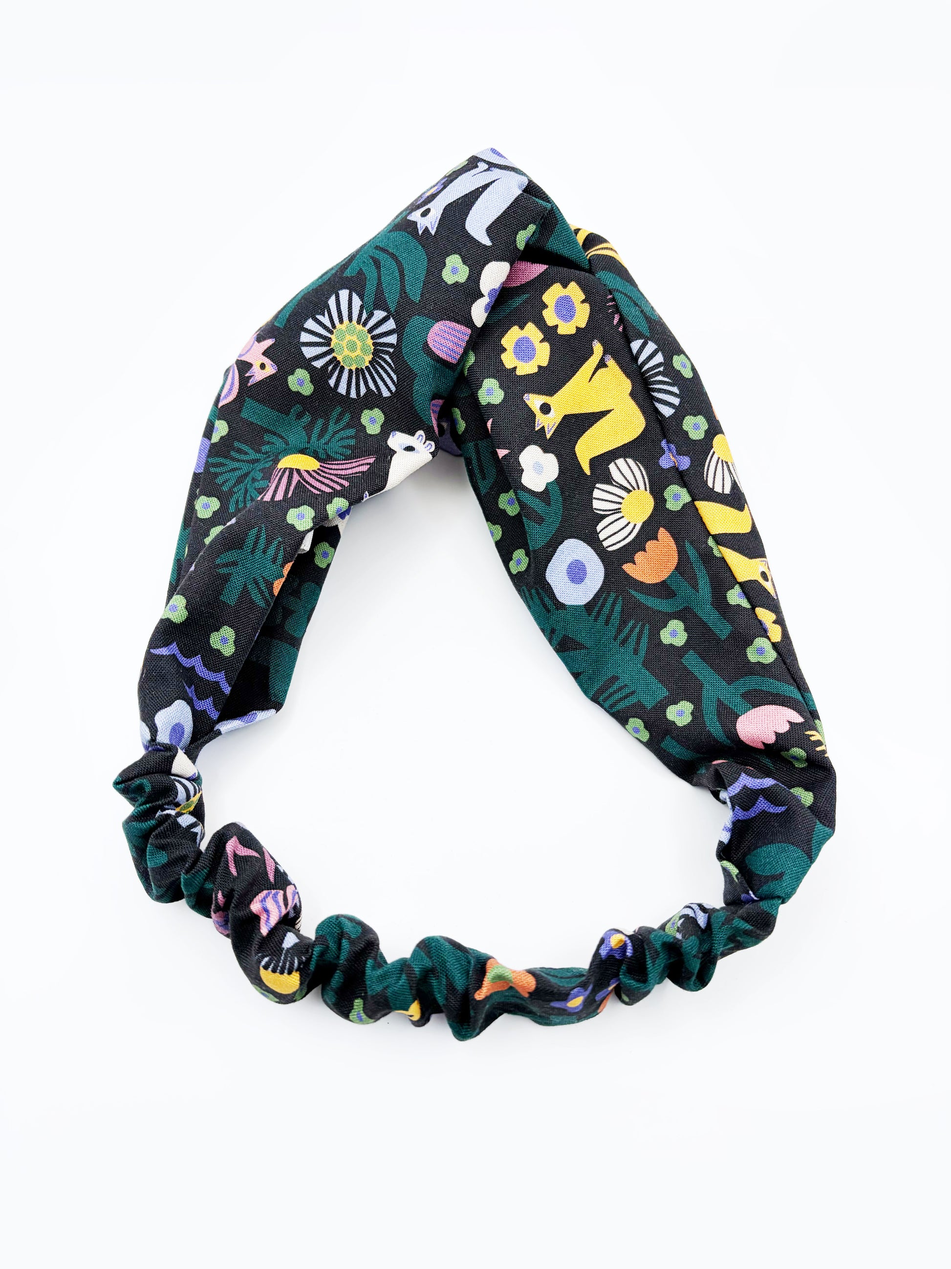 A black twist headband with a colorful abstract forest print featuring playful florals, leaves, and bright animals. The headband has an elastic band on the back.