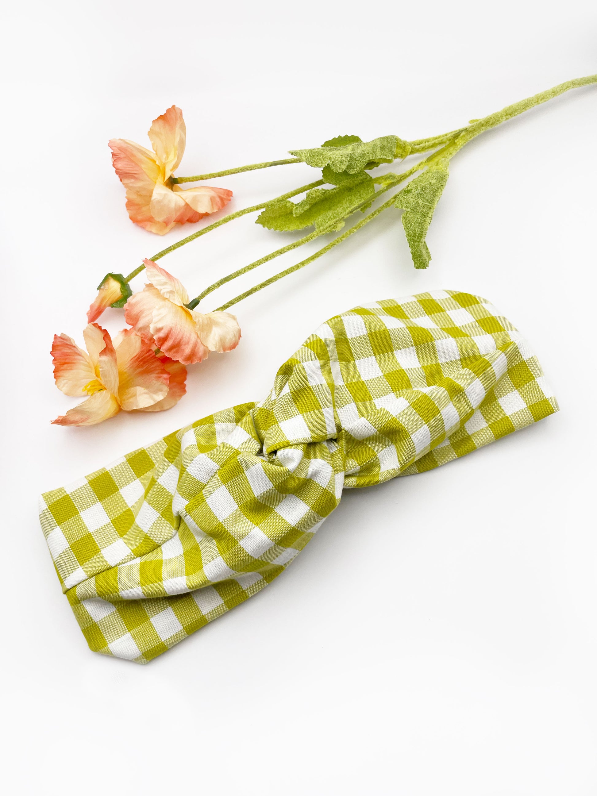 A handmade twist headband with a green and white gingham plaid print. The headband has an elastic band in the back. It is next to some pink artificial flowers.