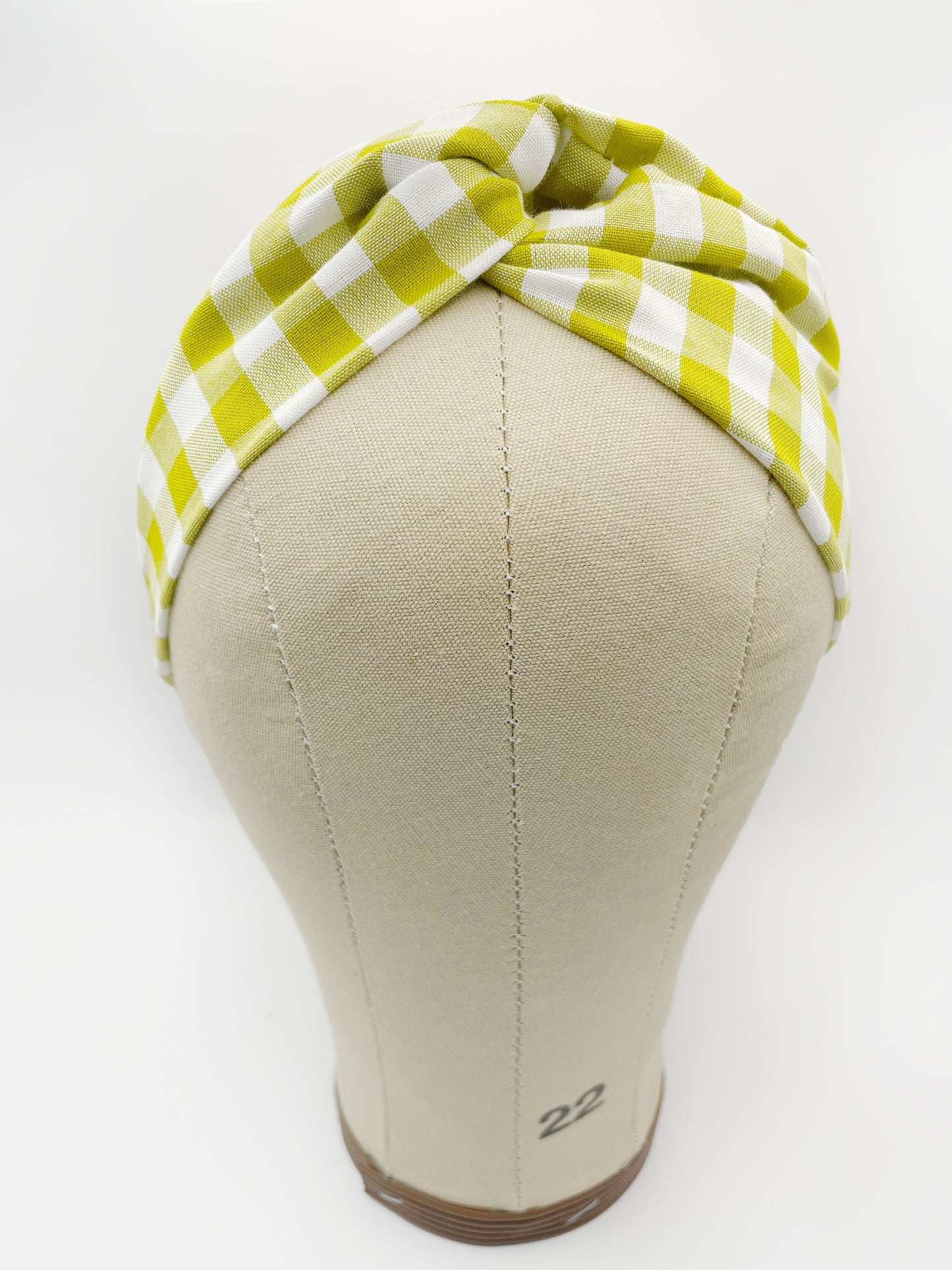 A handmade twist headband with a green and white gingham plaid print. The headband has an elastic band in the back. It is modeled on a mannequin head.