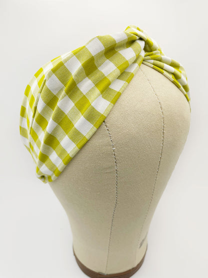 A handmade twist headband with a green and white gingham plaid print. The headband has an elastic band in the back. It is modeled on a mannequin head.