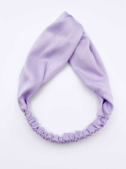 A twist headband made with a light purple, lavender crinkle satin fabric. The headband has an elastic band in the back.