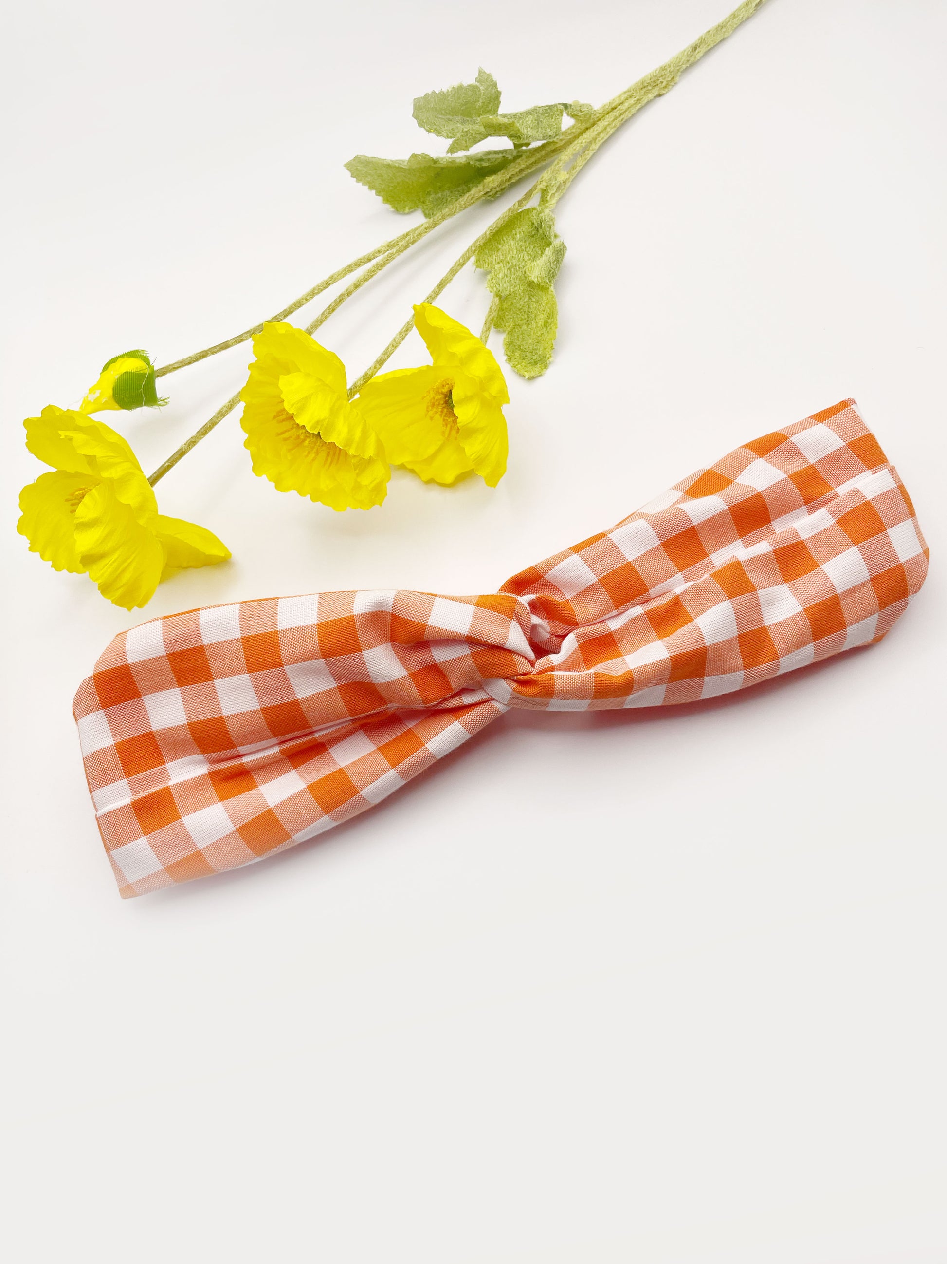 A handmade orange and white gingham plaid twist headband with an elastic band in the back. The headband is next to yellow artificial flowers.