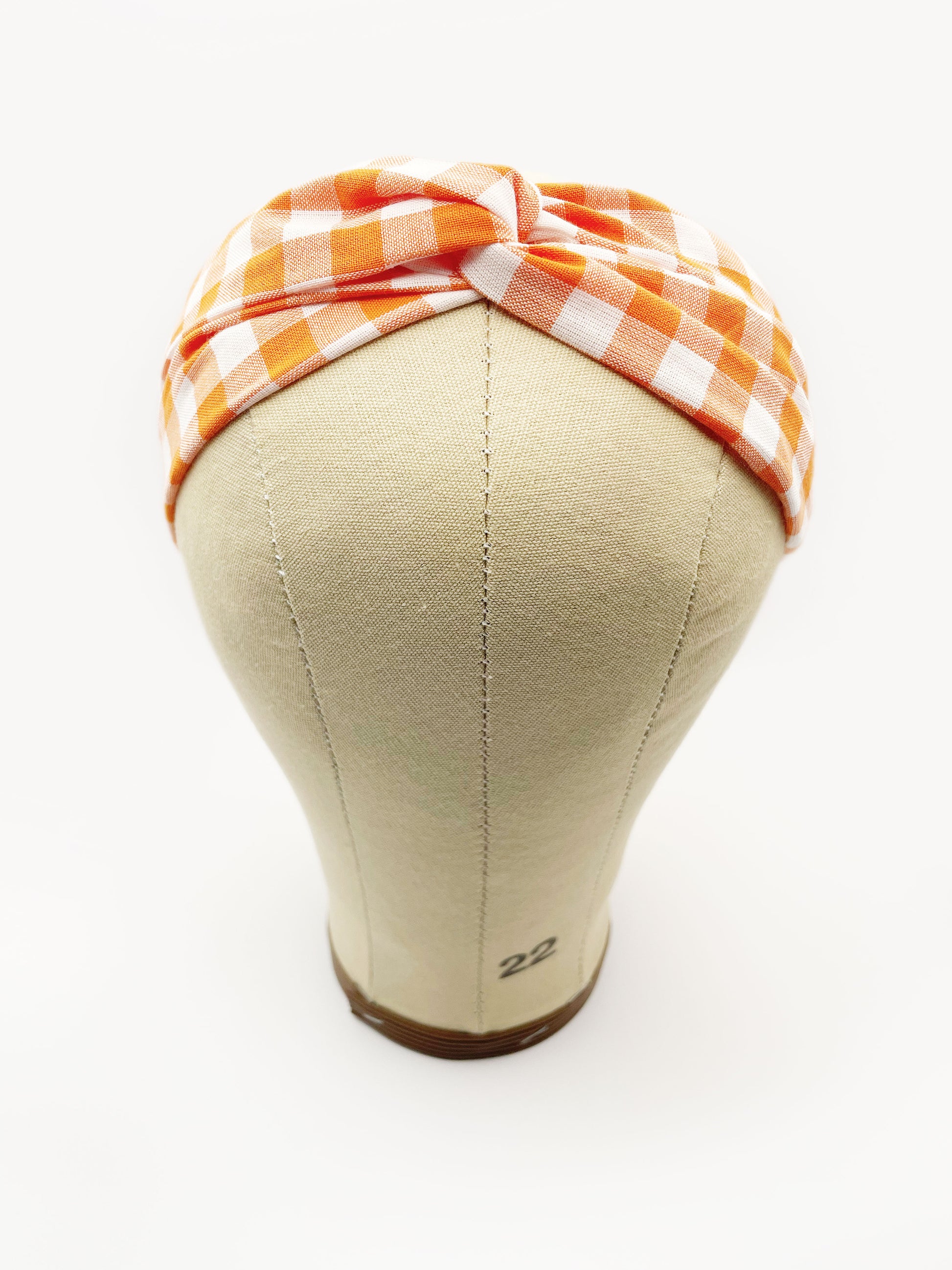 A handmade orange and white gingham plaid twist headband with an elastic band in the back. The headband is modeled on a mannequin head.