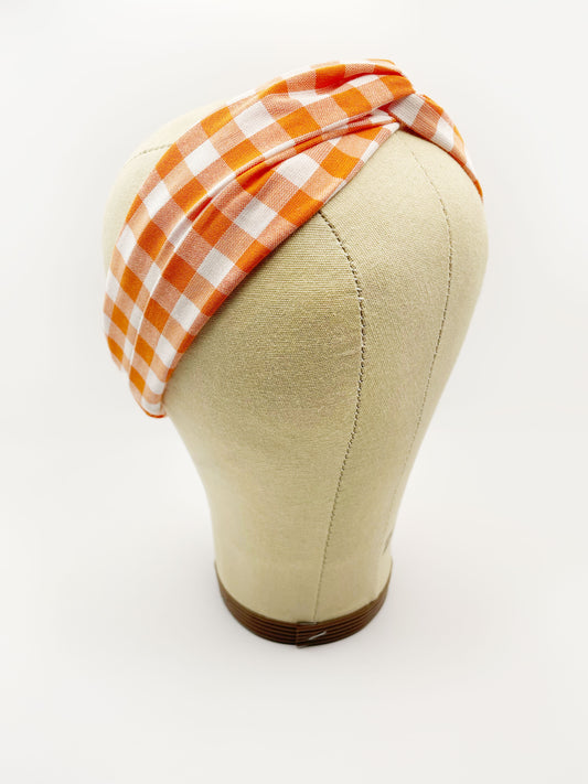 A handmade orange and white gingham plaid twist headband with an elastic band in the back. The headband is modeled on a mannequin head.