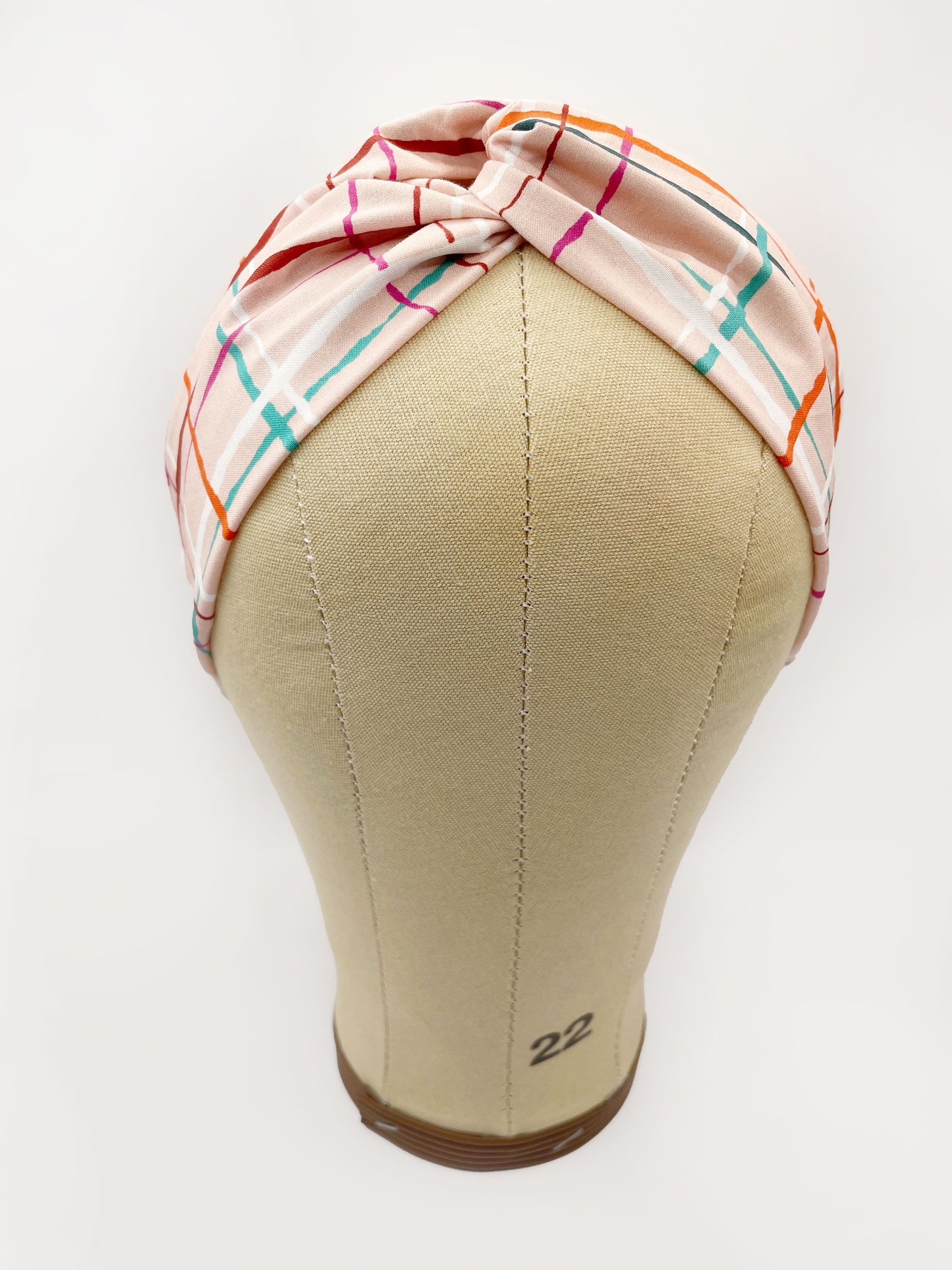 A fabric headband featuring a colorful and modern plaid design with vibrant shades of pink, red, teal, and orange on a soft pastel background. The headband is shown on a mannequin head.