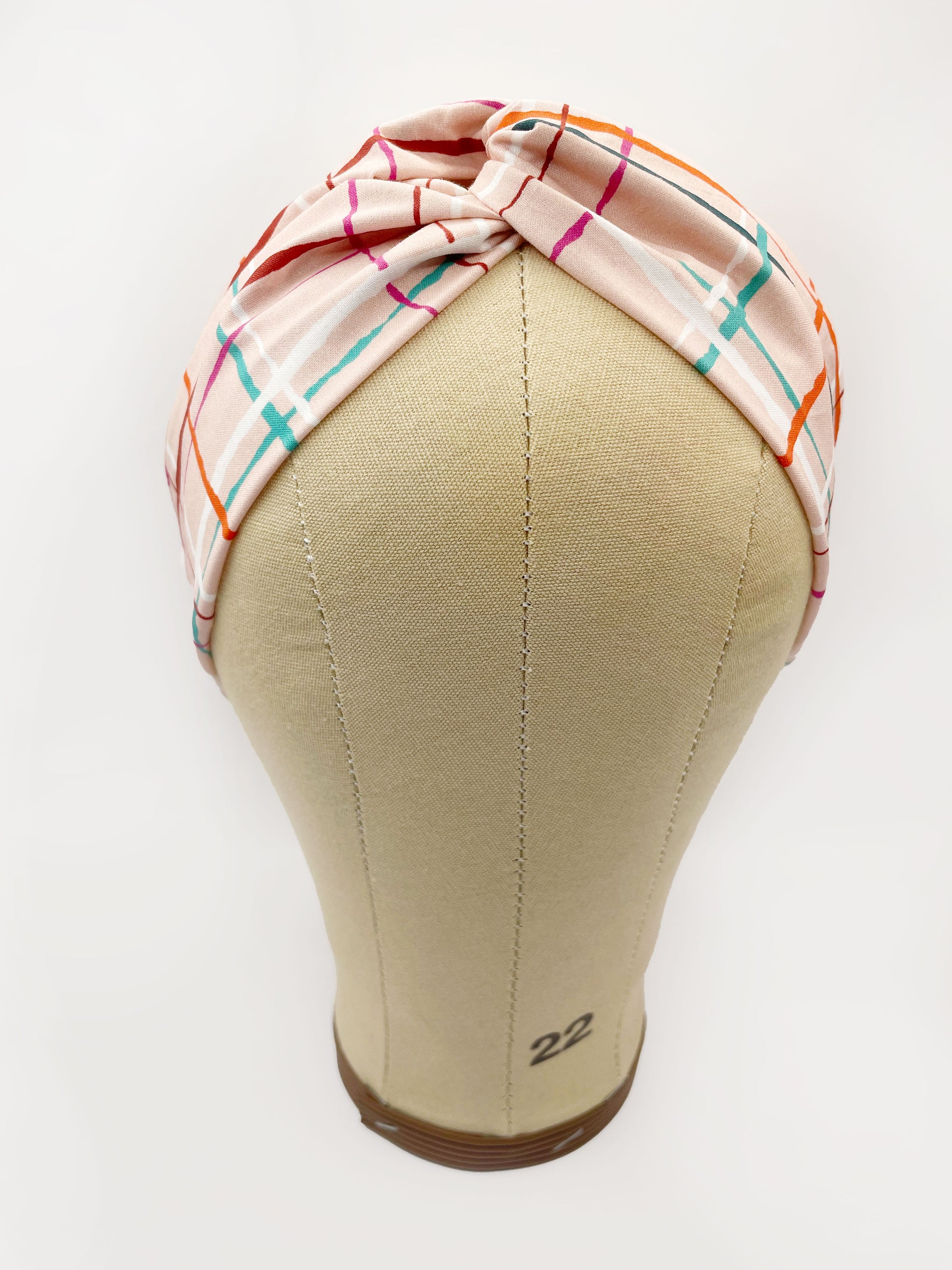 A fabric headband featuring a colorful and modern plaid design with vibrant shades of pink, red, teal, and orange on a soft pastel background. The headband is shown on a mannequin head.